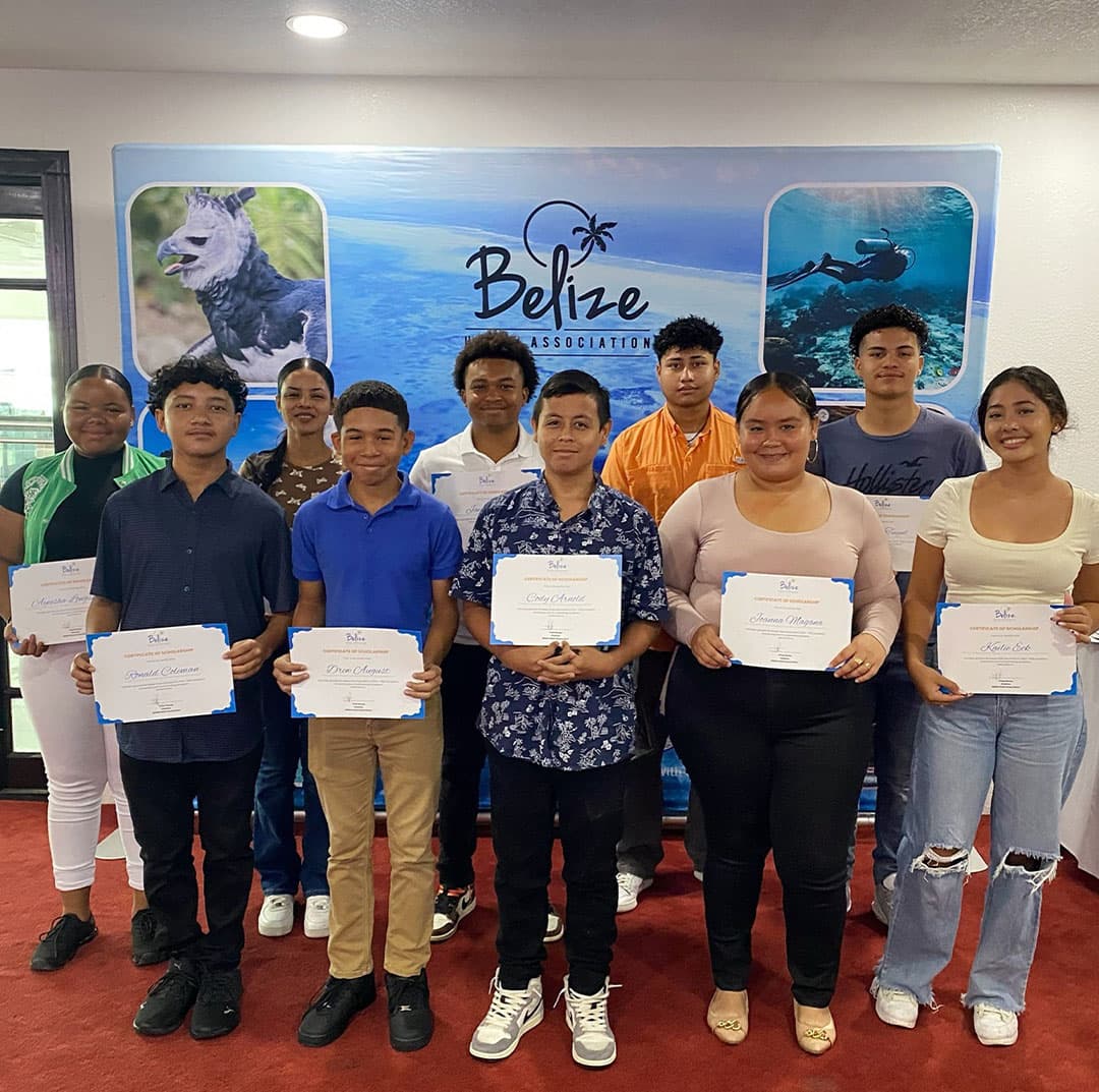 A group of students proudly holding certificates in front of a Belize Association backdrop.