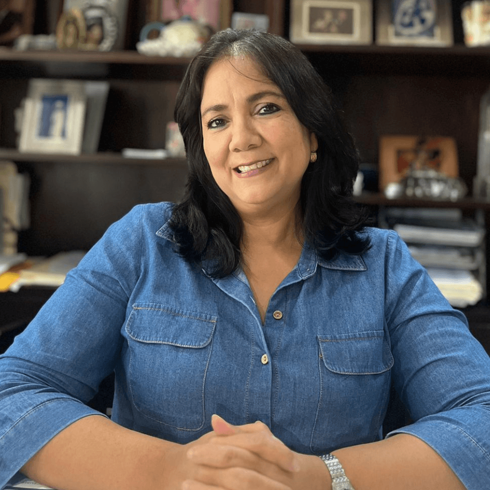 A woman with dark hair smiles while seated at a desk, wearing a denim shirt.