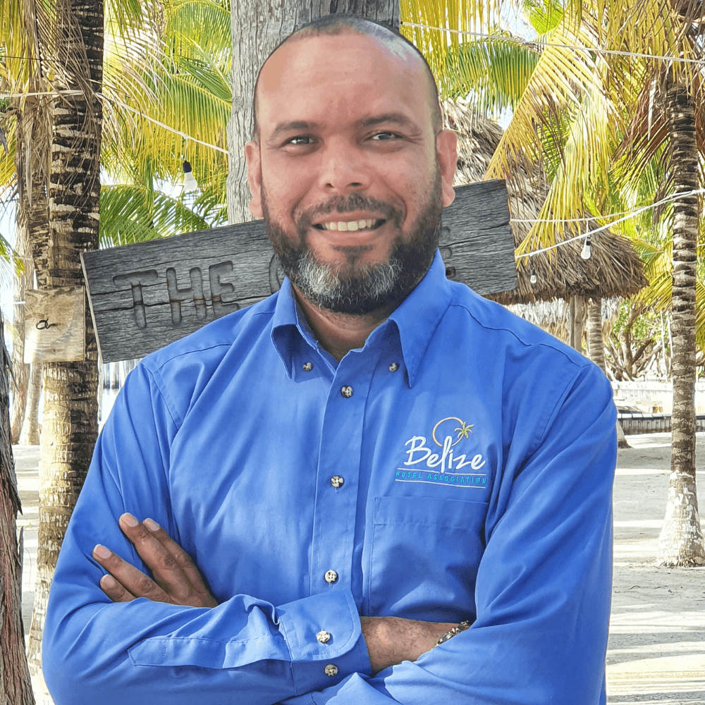 A man in a blue shirt with "Belize Hotel Association" logo stands confidently with arms crossed in a tropical setting.