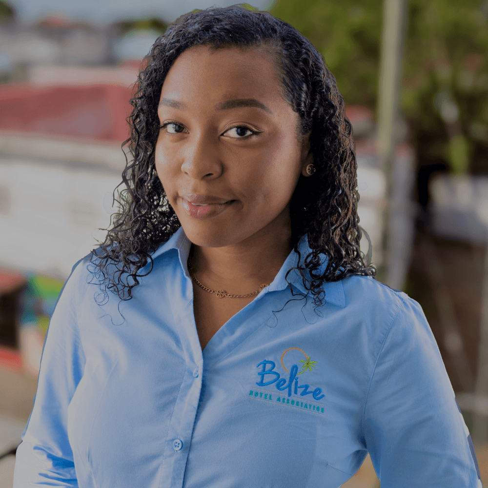 A woman with curly hair smiles confidently while wearing a light blue shirt featuring the logo of the Belize Hotel Association.