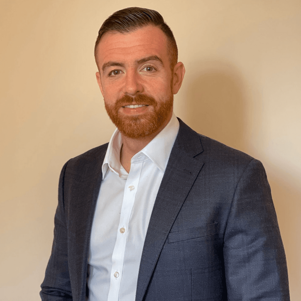 A man with a neatly trimmed beard and dressed in a suit smiles against a neutral background.