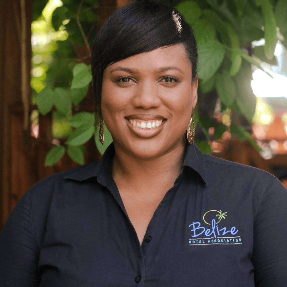 A smiling woman wearing a black shirt with the Belize Hotel Association logo stands in front of lush green foliage.