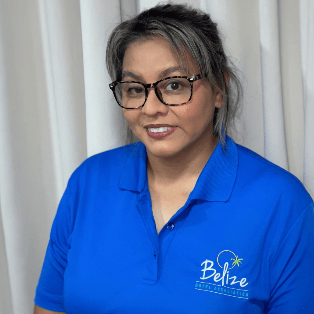 A woman in a blue polo shirt with the Belize Hotel Association logo stands in front of a white curtain.