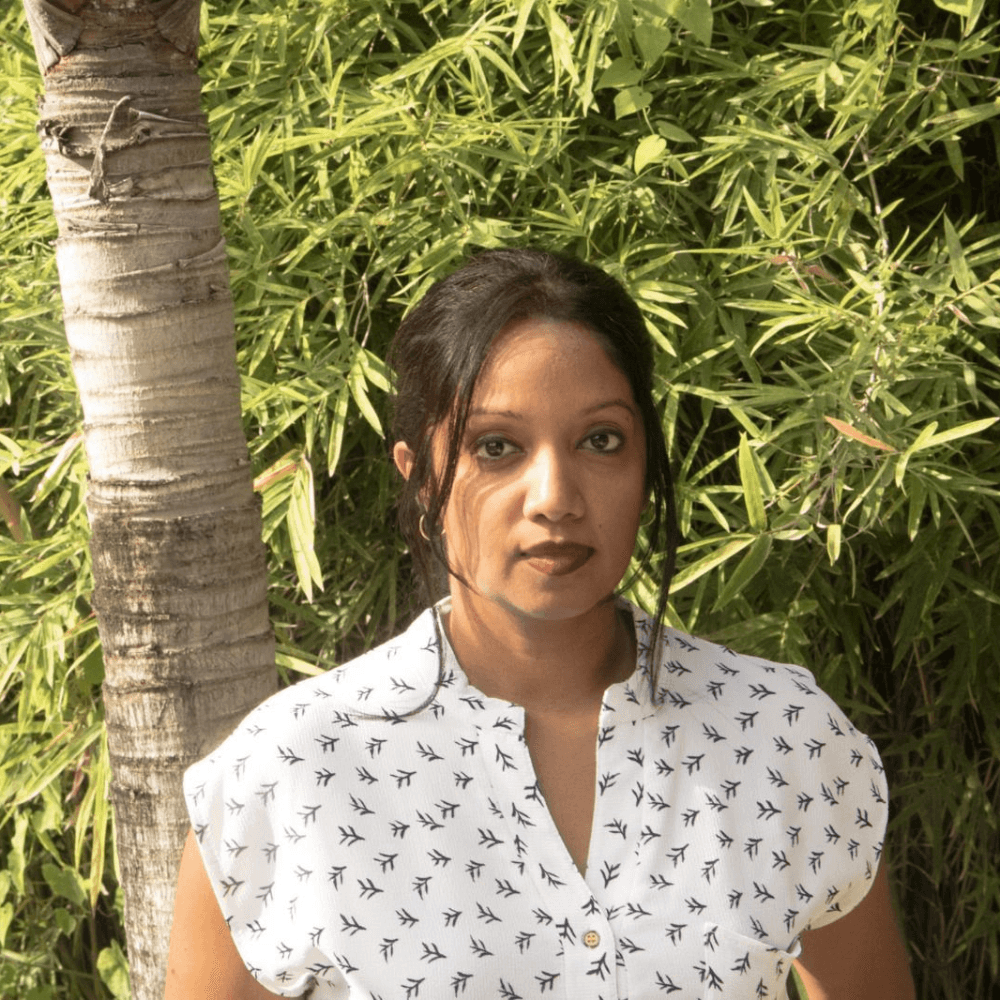 A woman with long black hair, wearing a white shirt with a pattern, stands in front of a green bamboo background.