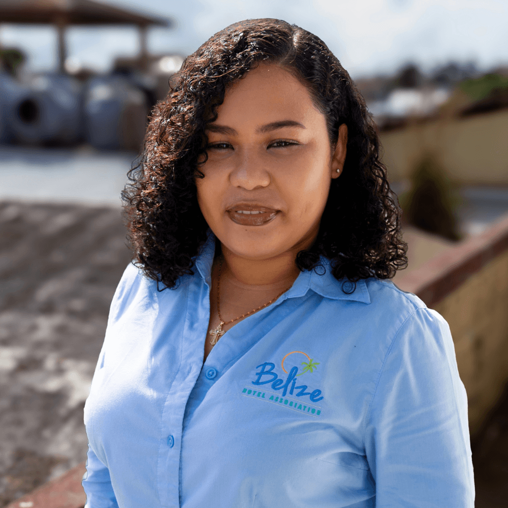 A woman with curly hair wearing a blue shirt with a "Belize Hotel Association" logo stands outdoors.
