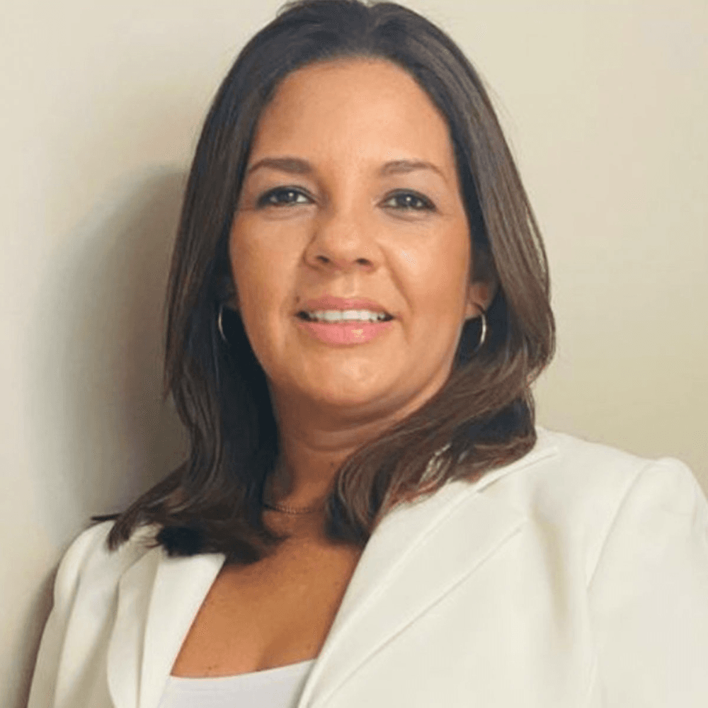 A woman with shoulder-length hair smiles while wearing a white blazer against a neutral background.
