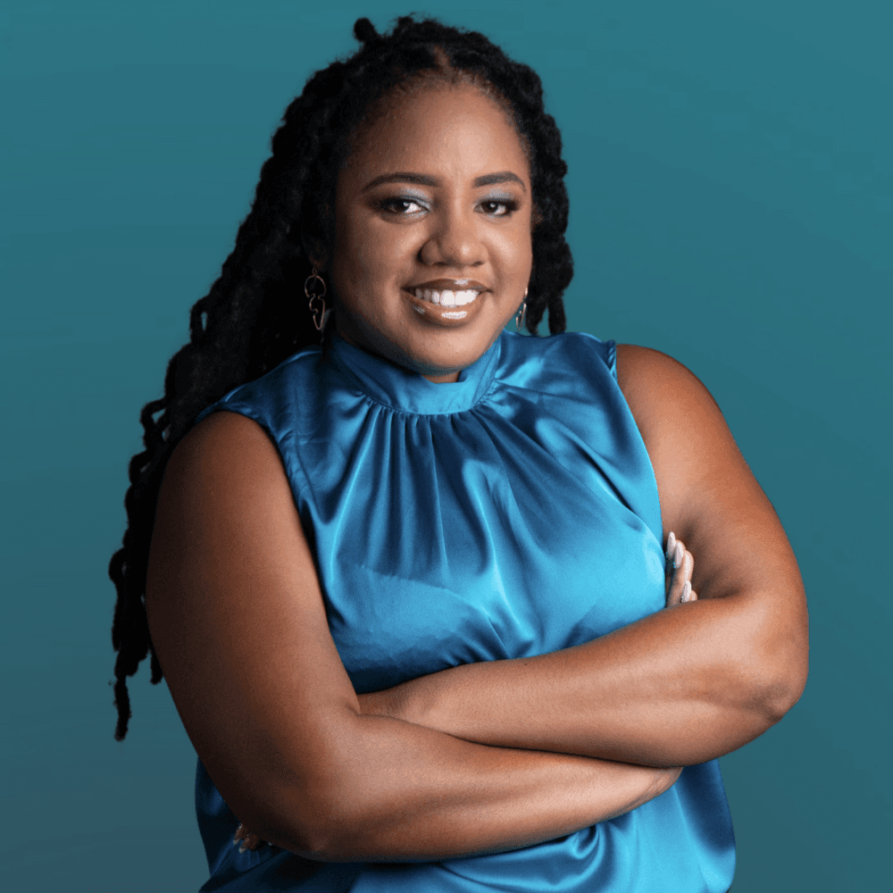 A smiling woman with long, textured hair crosses her arms while wearing a teal blouse against a solid background.