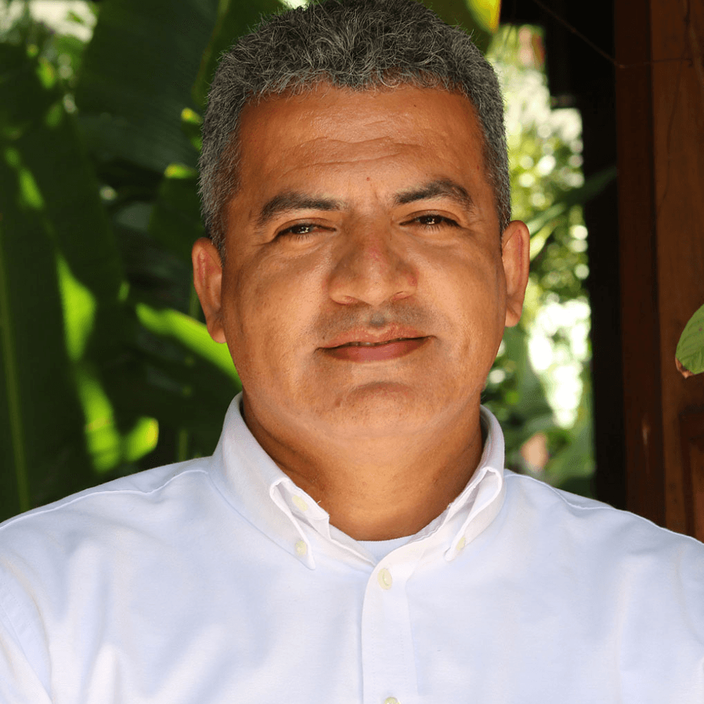 A man in a white shirt smiles against a backdrop of green foliage.