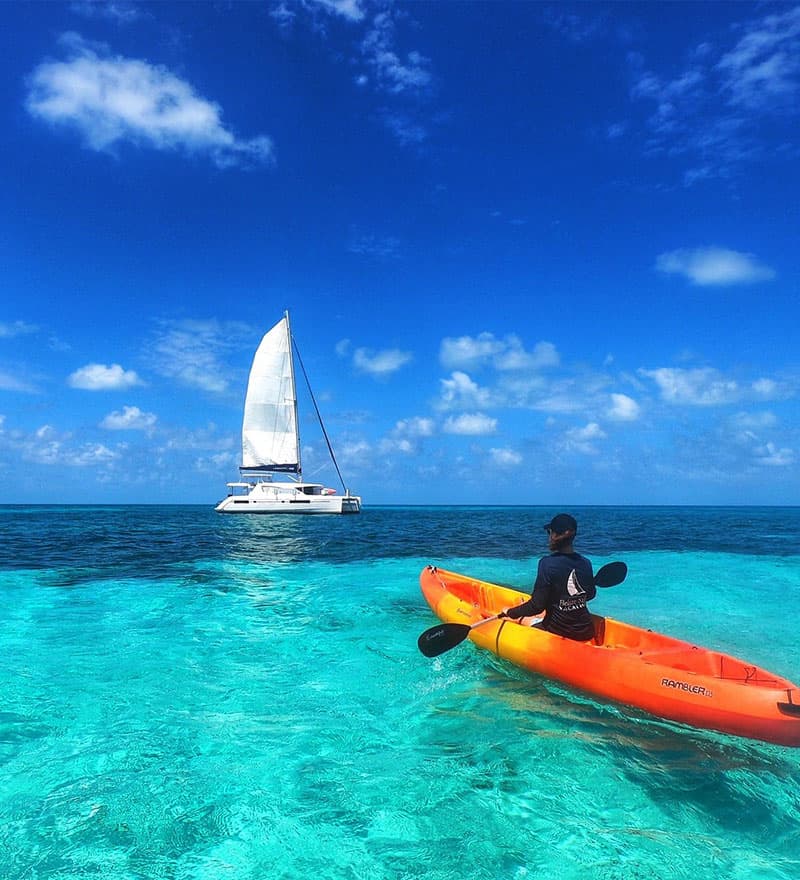 A person paddles a bright orange kayak on clear blue water with a sailboat in the background.