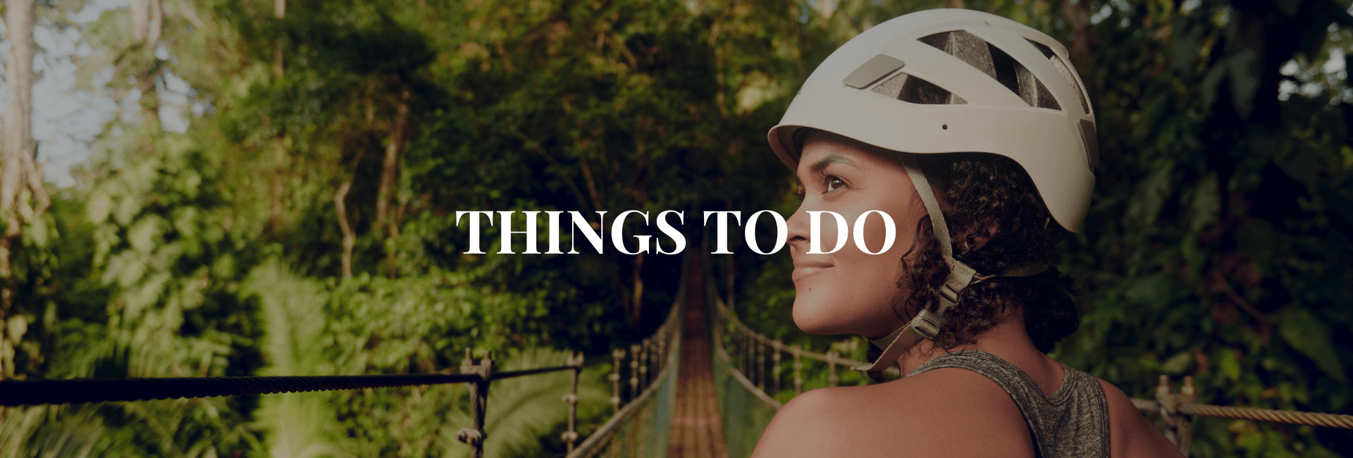A woman wearing a helmet smiles while standing on a bridge surrounded by lush greenery.