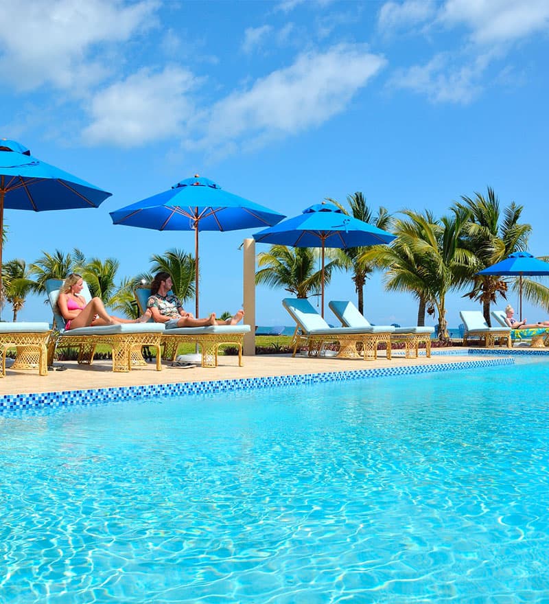 Two women relax by a blue pool under sunny skies and bright umbrellas.