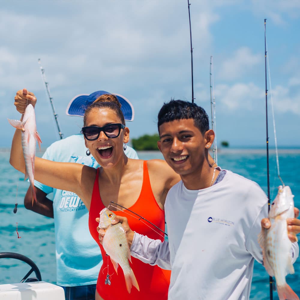 A smiling woman in a red swimsuit and a man hold caught fish while fishing on a boat under a blue sky.