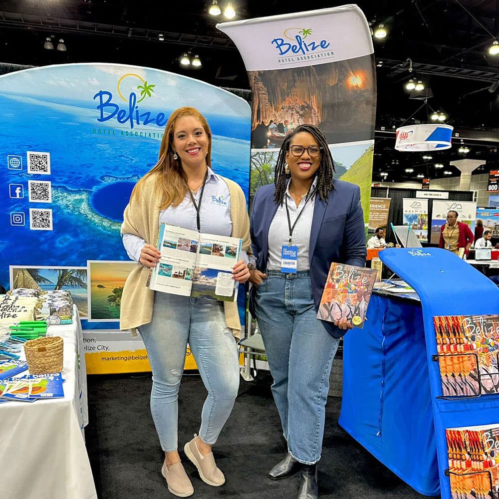 Two women stand at a booth promoting Belize tourism, holding brochures amidst colorful displays.